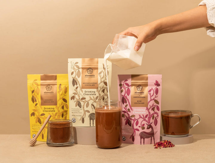 Person pouring milk into a glass of hot chocolate with various chocolate products on a beige background