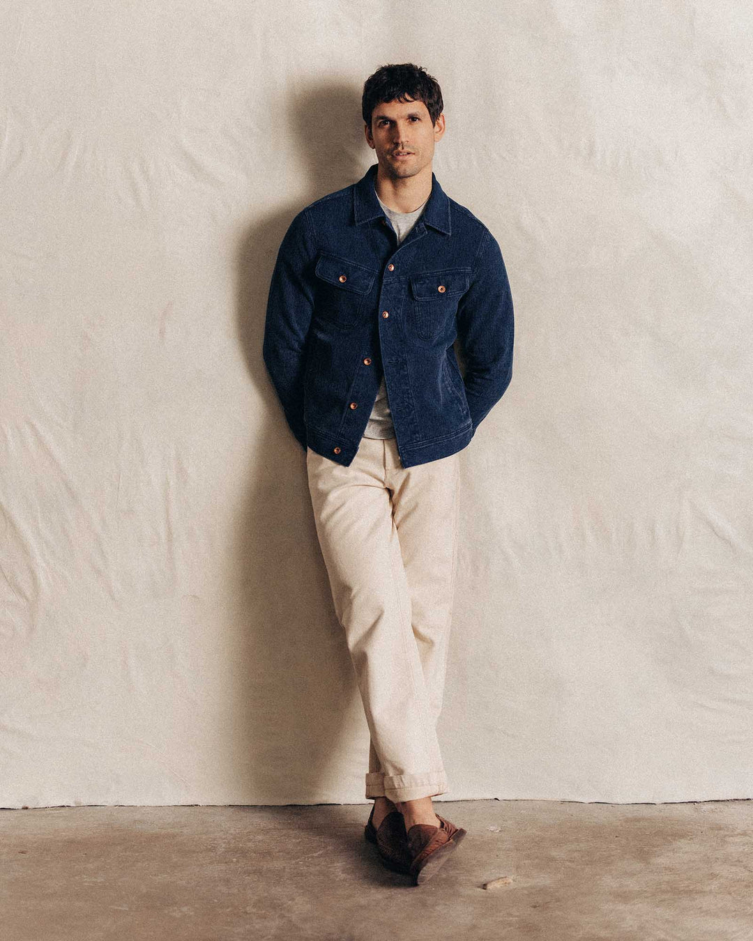 man wearing jean jacket and white pants against a white wall.