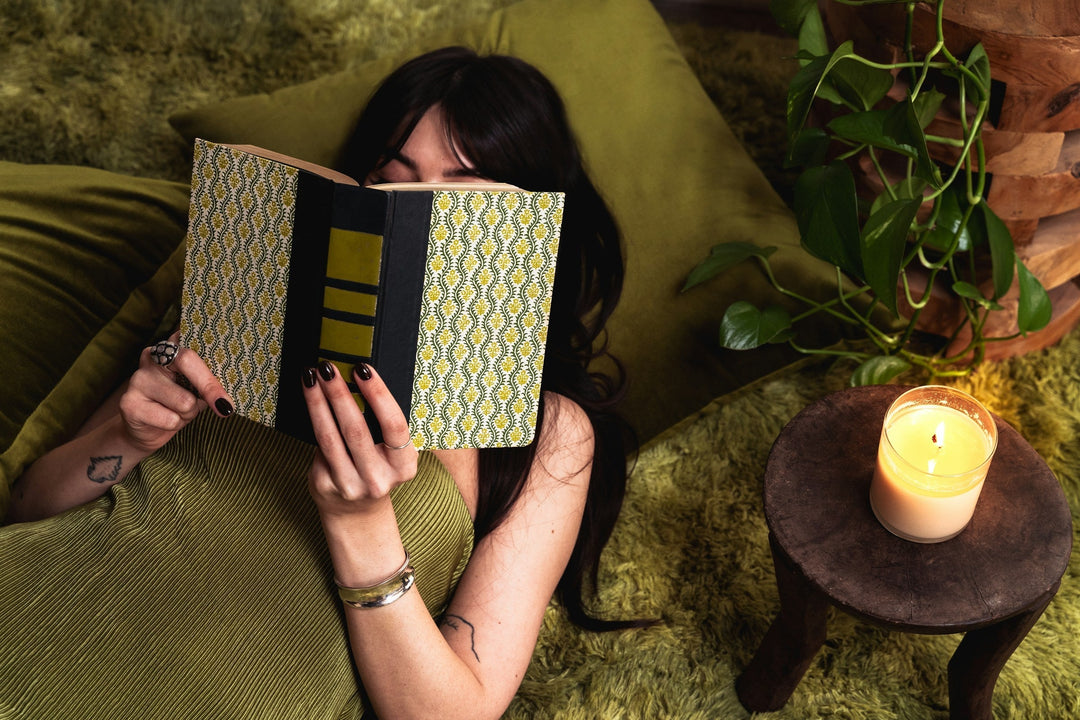 A dark-haired woman wearing a green dress reading a book by candlelight
