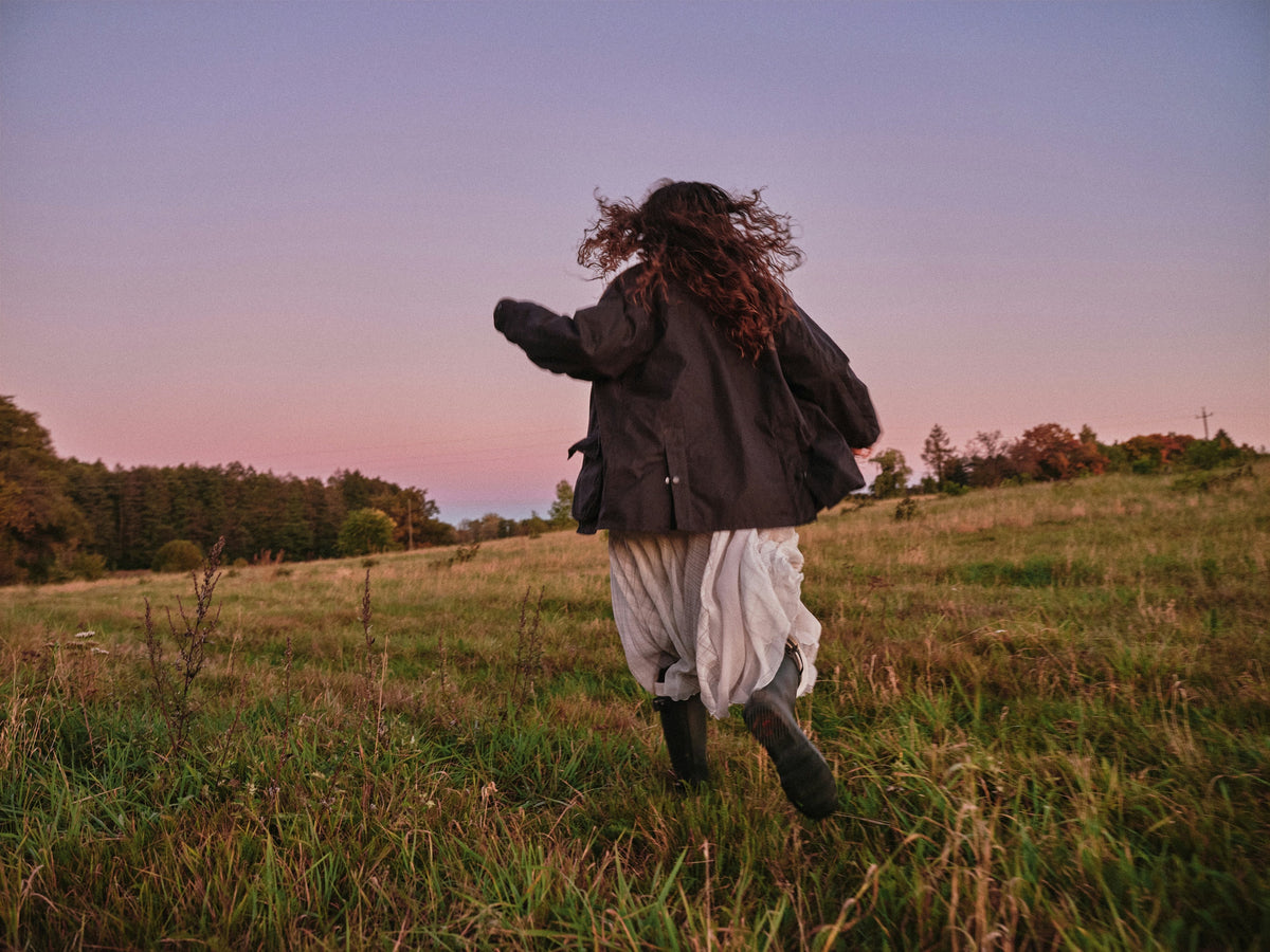 A person wearing an oversized jacket, white skirt, and boots, running through a field at dusk