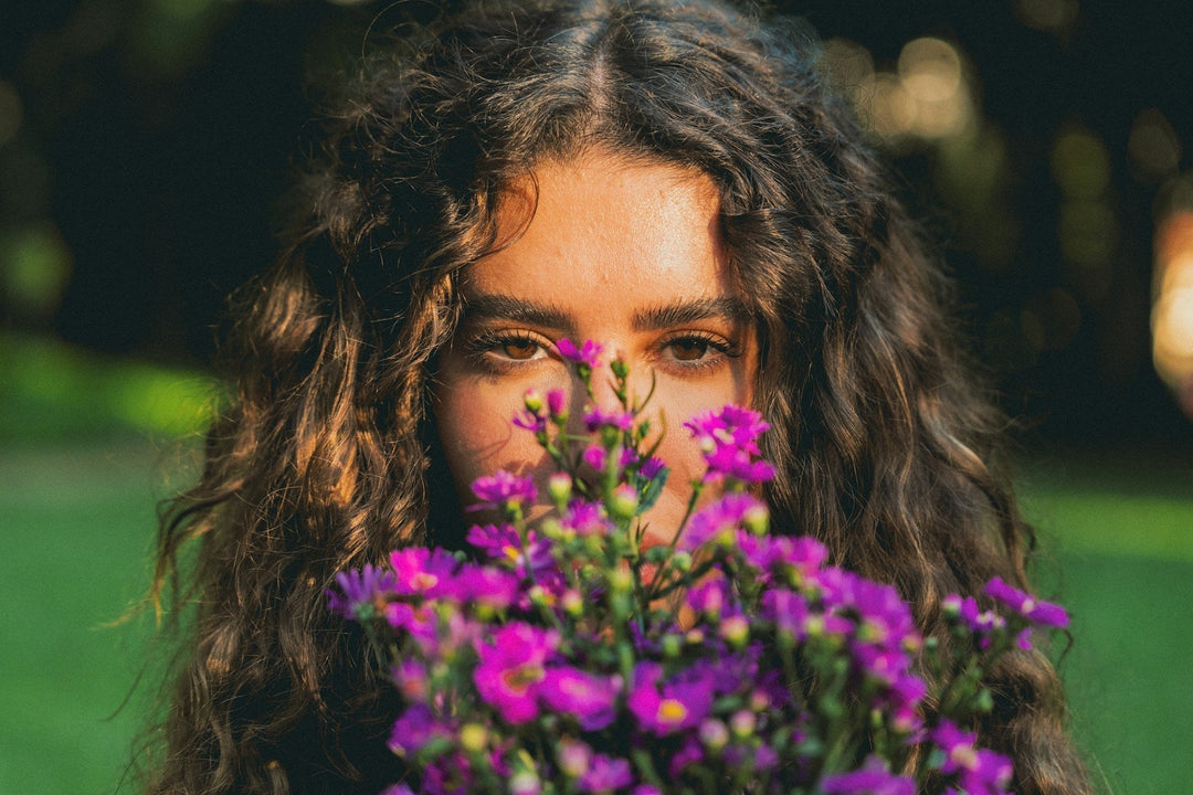 A dark-haired woman holding brilliant purple flowers in front of her face during golden hour