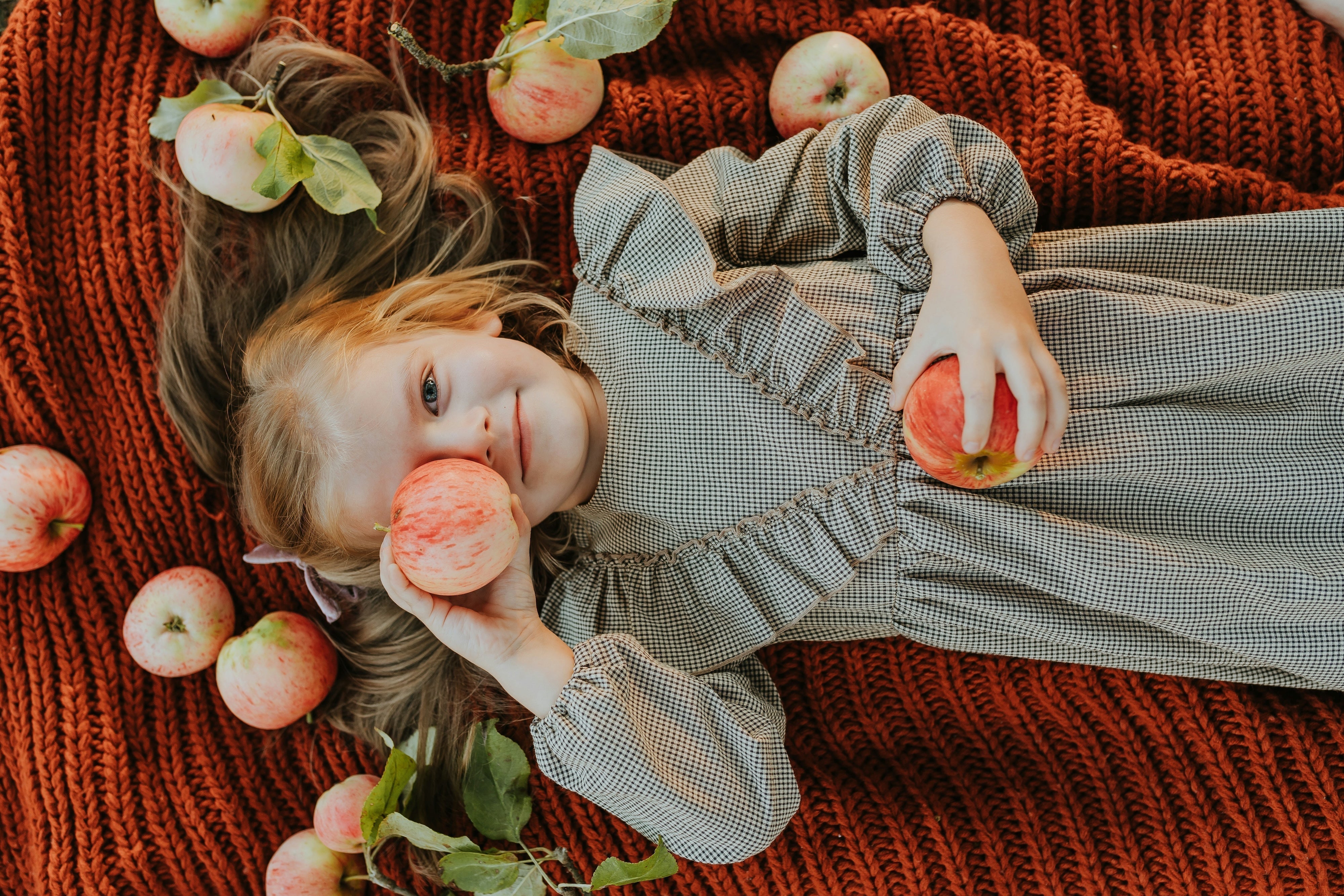 A young girl laying on an orange knit blanket holding an apple over her right eye