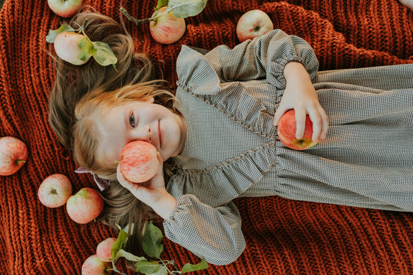 A young girl laying on an orange knit blanket holding an apple over her right eye