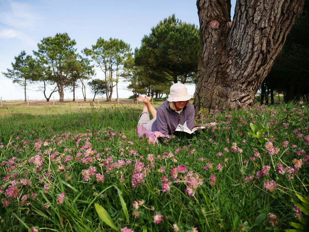 A person wearing a white bucket hat sitting beside a tree in a field of green grass and pink flowers