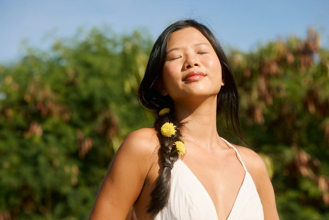 A smiling, dark-haired woman standing with eyes closed facing the sun with flowers in her braided hair
