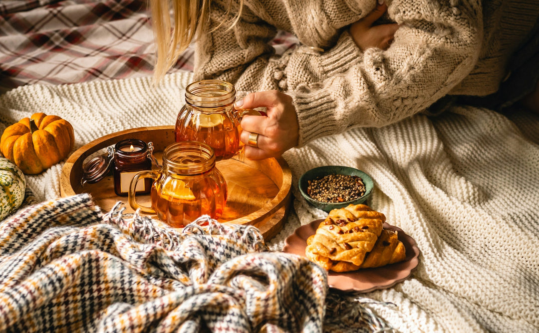 A person's arms visibly reaching towards two mugs of tea set on a tray upon on a bed of cozy blankets