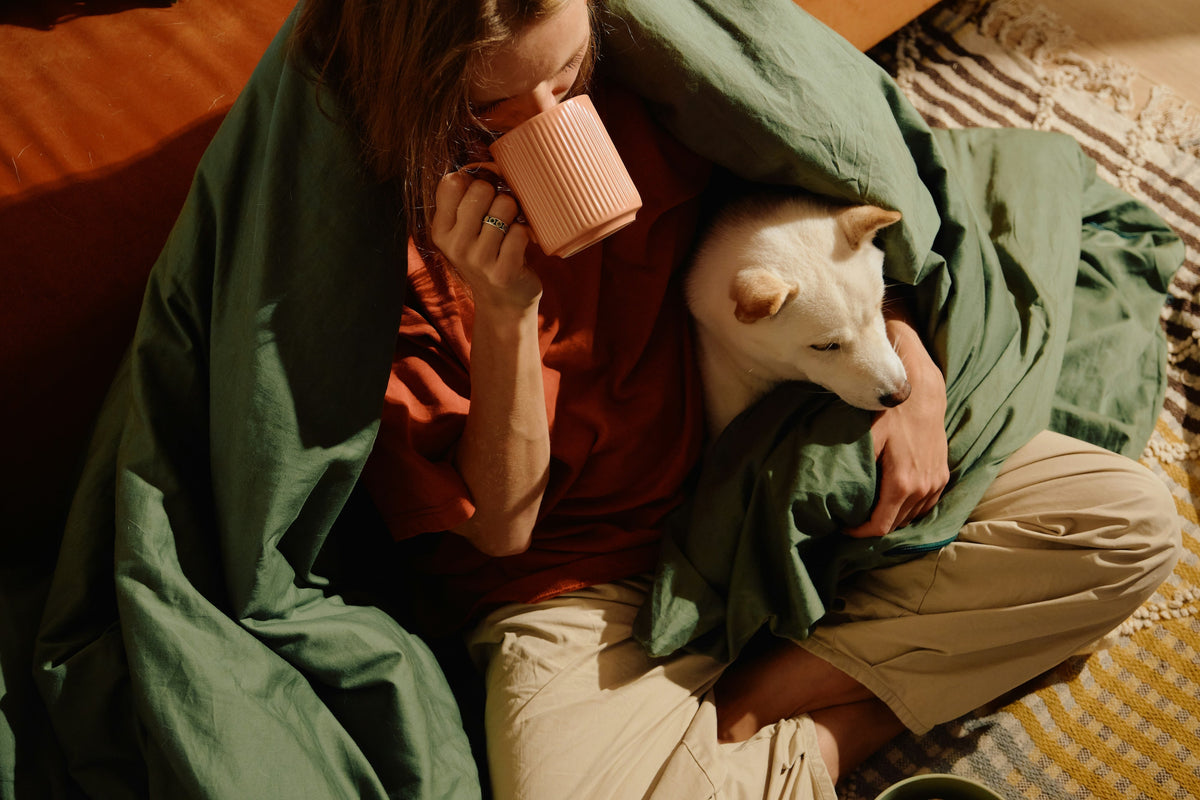 A person wrapped in a green comforter, drinking from a pink mug, while snuggling a dog