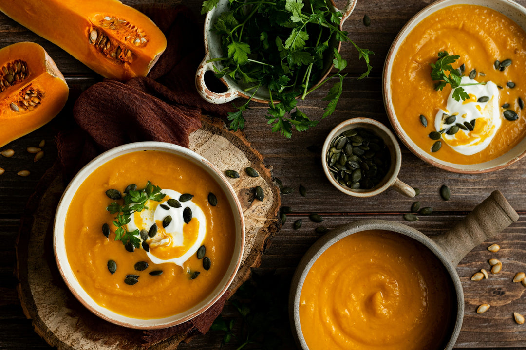 Three bowls of a vibrant orange squash soup, raw butternut squash, and a bowl of parsley