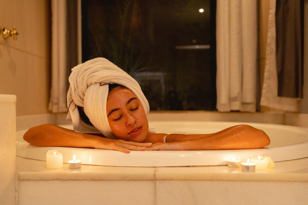 A person in the bath, wearing a plush white towel on her head to dry their hair, with candles lit on the bathtub edge.
