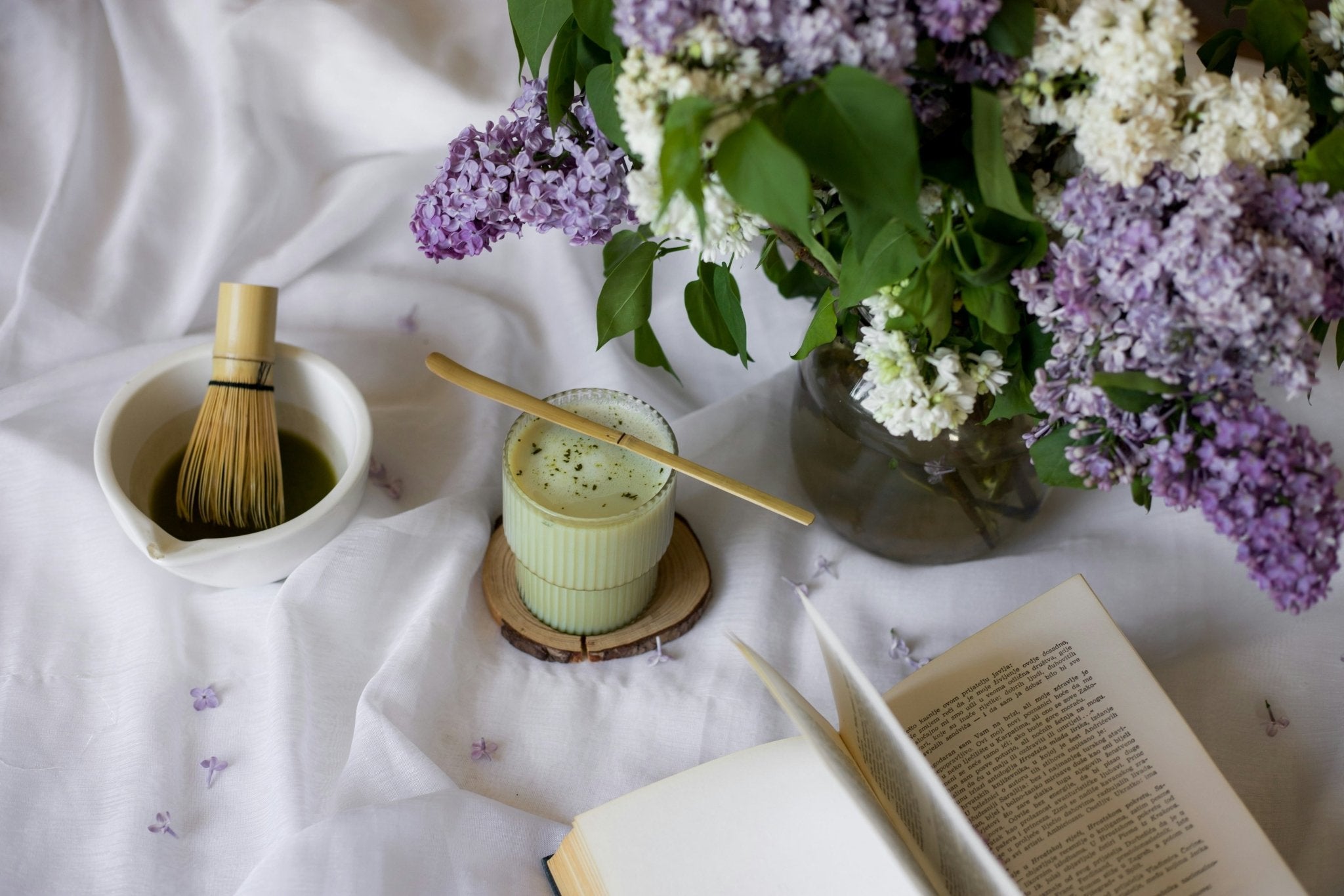 Iced matcha set on a white fabric background next to a vase of lilacs and an open book