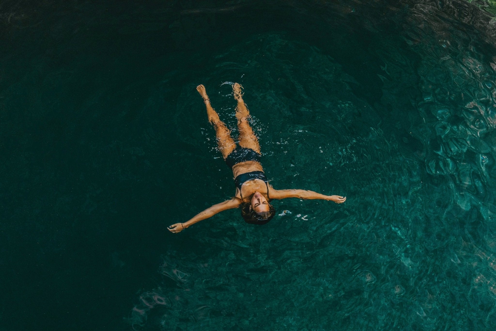 A woman floating peacefully in teal waters wearing a black swimsuit