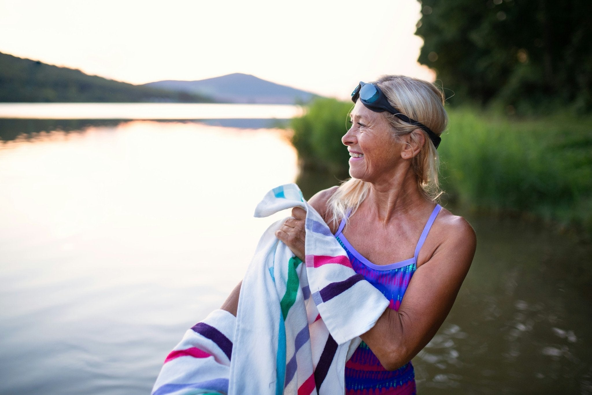 A mature woman smiling, wearing a bathing suit and swimming goggles, standing in a river at golden hour