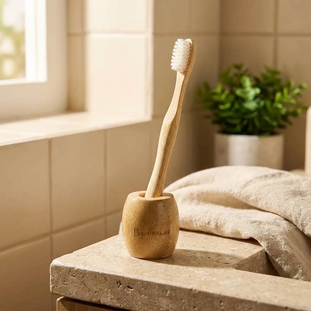 Bamboo toothbrush in a wooden holder on a bathroom counter with a towel and plant in the background.
