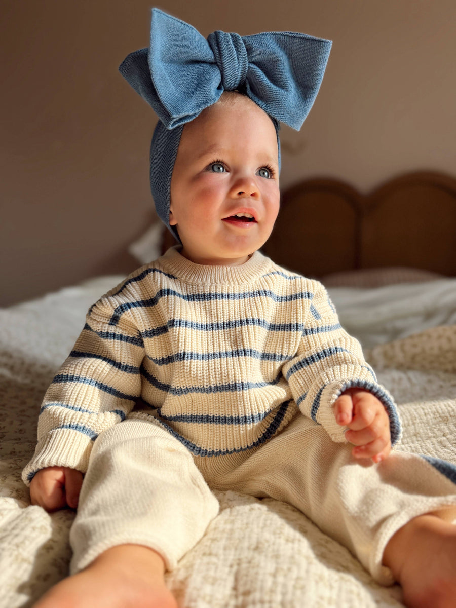 Baby wearing a blue striped sweater and headband sitting on a bed.