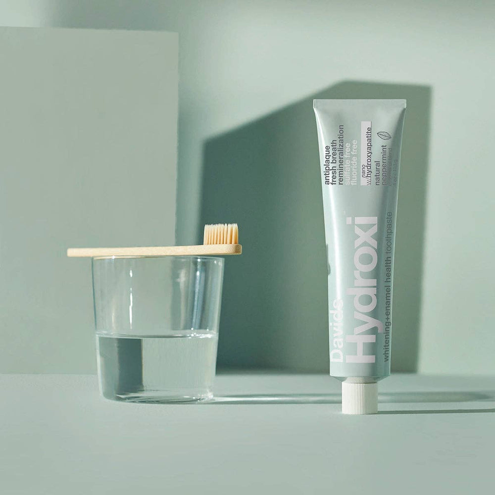Tube of Davids Hydroxi toothpaste with a glass and toothbrush on a light gray background
