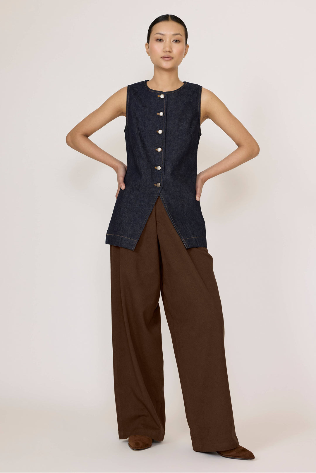Woman wearing a Laude Longline Vest in Dark Indigo with brown pants over a plain background