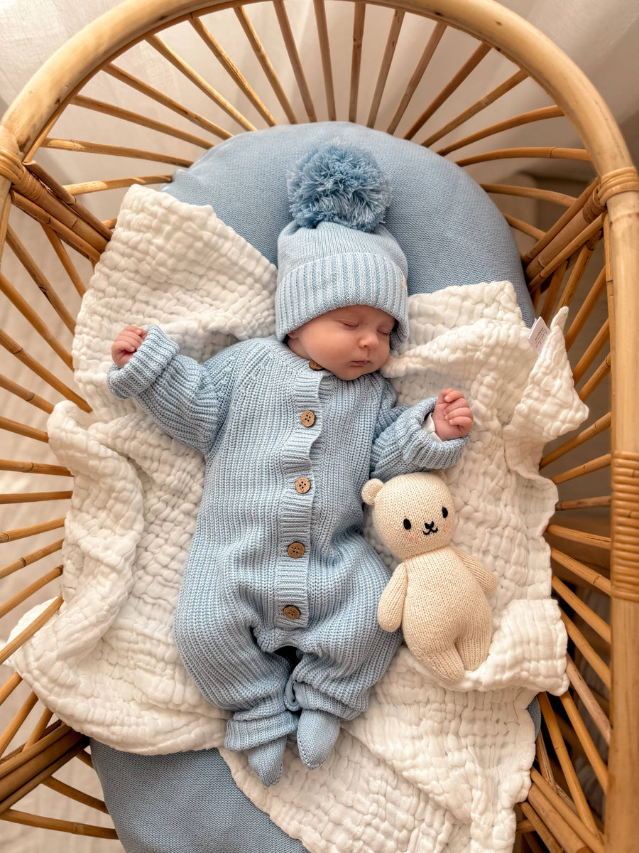 Newborn baby in a blue outfit with a matching hat, lying on a textured blanket in a wicker basket.