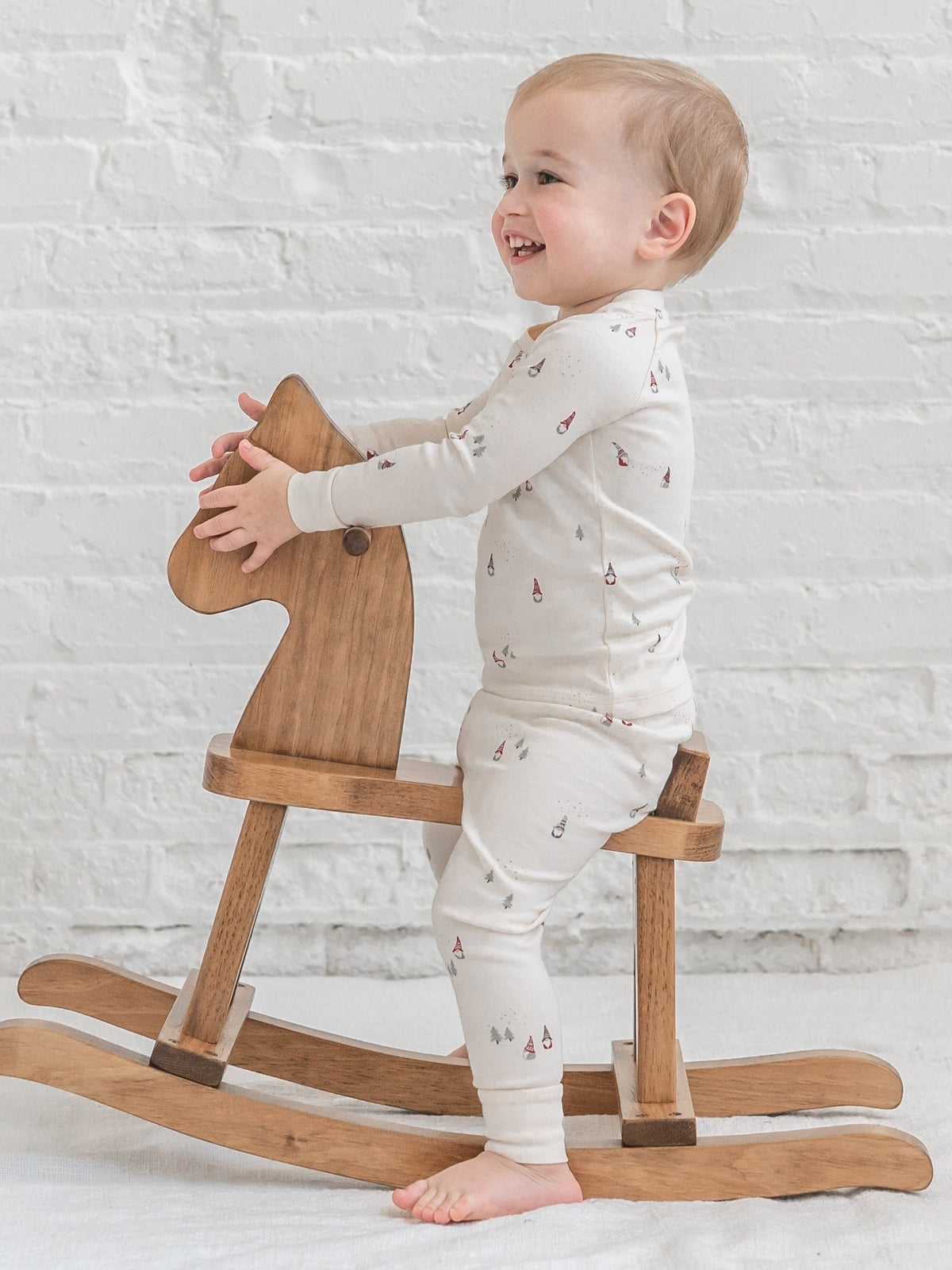 A baby standing on a wooden rocking horse, wearing a Colored Organics' 2 Piece Long Sleeve Jammie Set with a small red and green pattern. The background is a white brick wall.