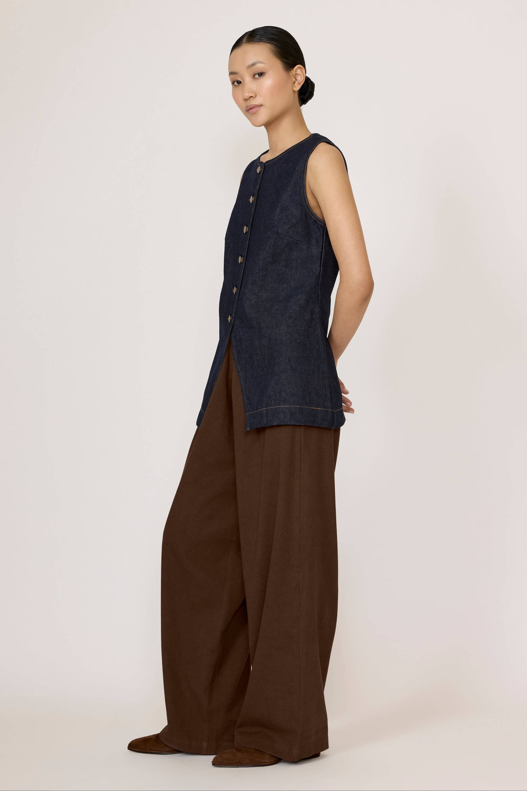 Woman wearing a Laude Longline Vest in Dark Indigo with brown pants over a plain background
