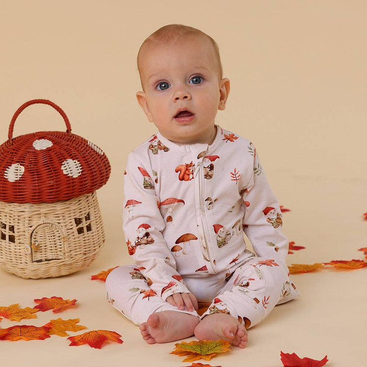 Baby wearing a Gnomes Organic Snugglesuit Convertible Romper, sitting next to a wicker basket with a red top on a beige background.