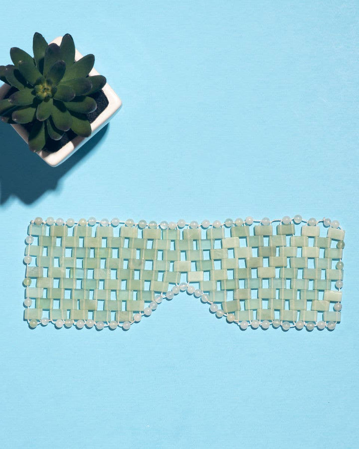 Green woven eye mask on a blue background with a small potted plant.