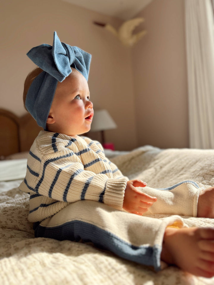 Baby wearing a blue headband and striped sweater sitting on a bed.