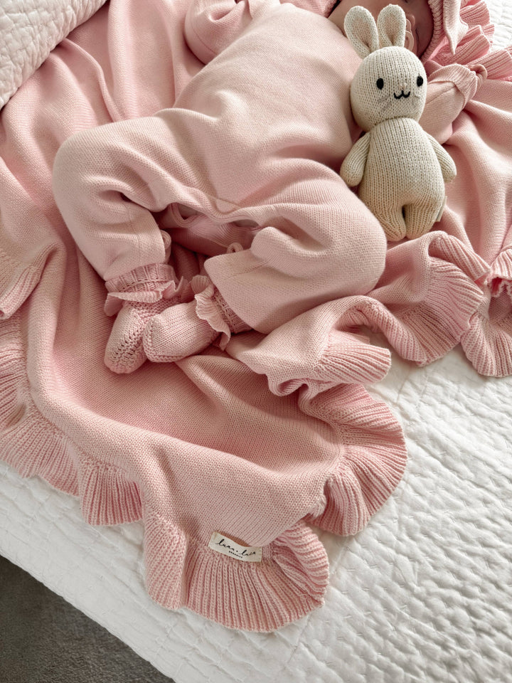 Pink baby outfit with matching booties and ruffled details and a soft toy on a textured surface