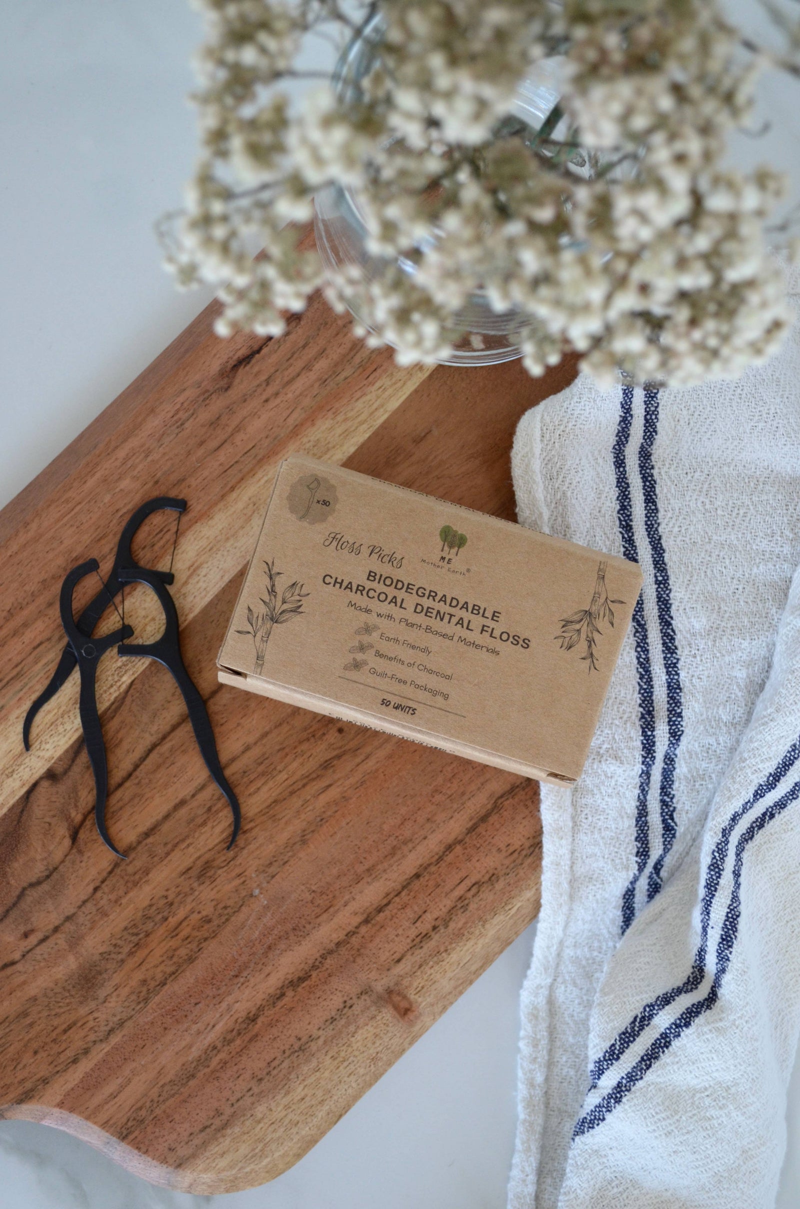 Biodegradable charcoal dental floss packaging on a wooden board with a towel and flowers in the background.