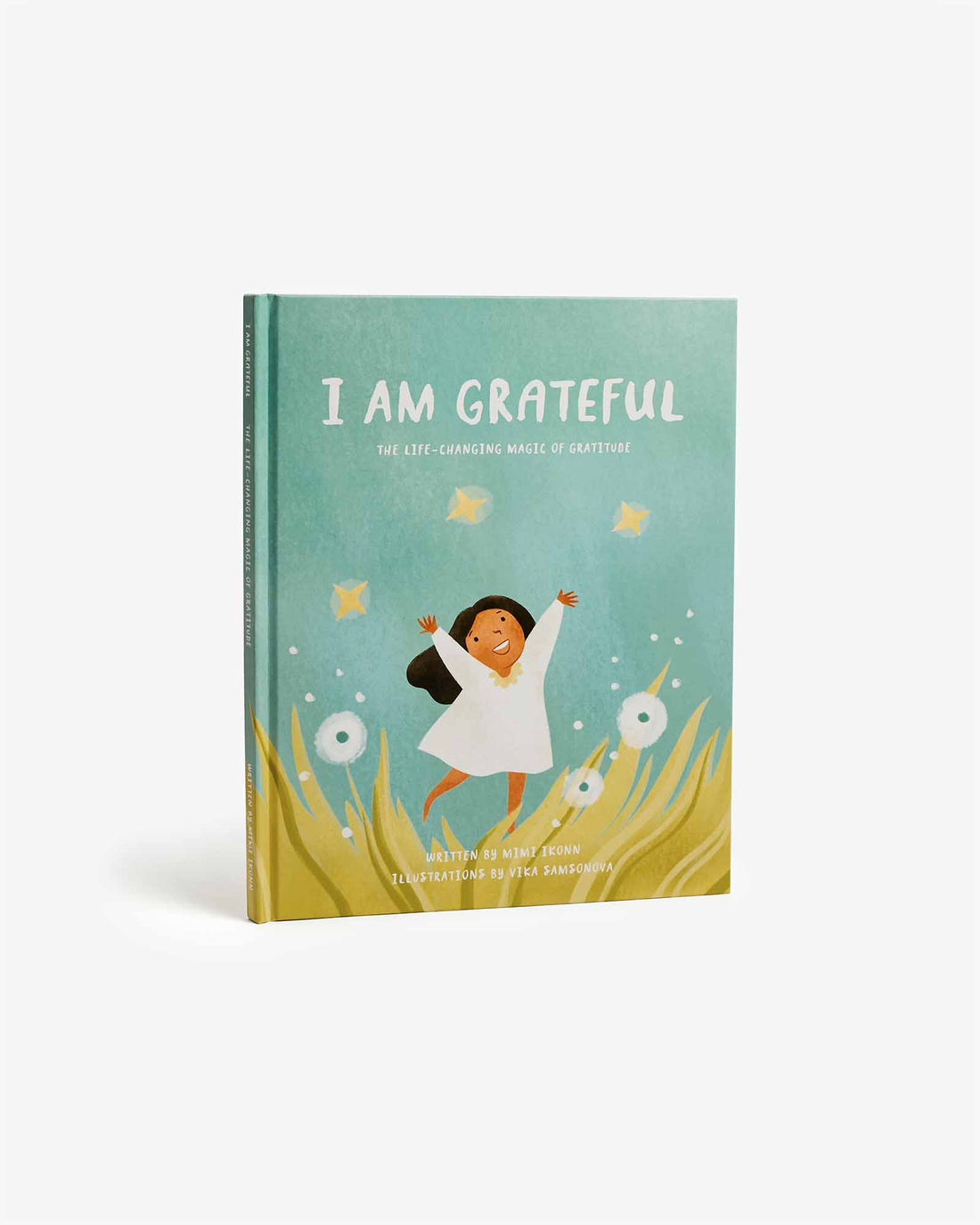 A children's book titled 'I AM GRATEFUL' shown standing upright against a clean white background. The cover features a teal background with an illustration of a smiling girl in a white dress jumping among yellow grass, with small white flowers and floating yellow shapes.