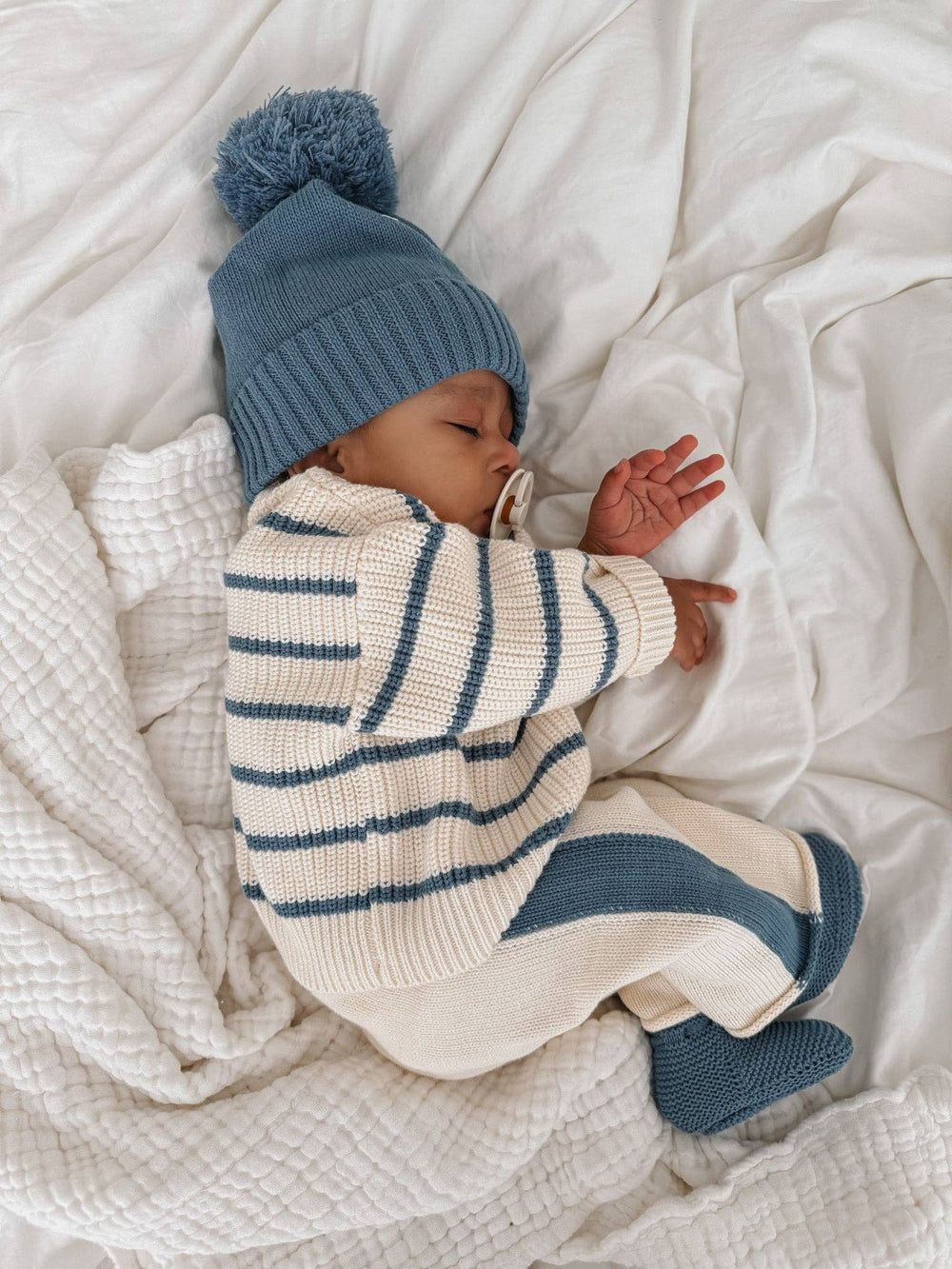 Baby wearing a blue and white striped outfit with a matching hat and socks, lying on a white blanket.