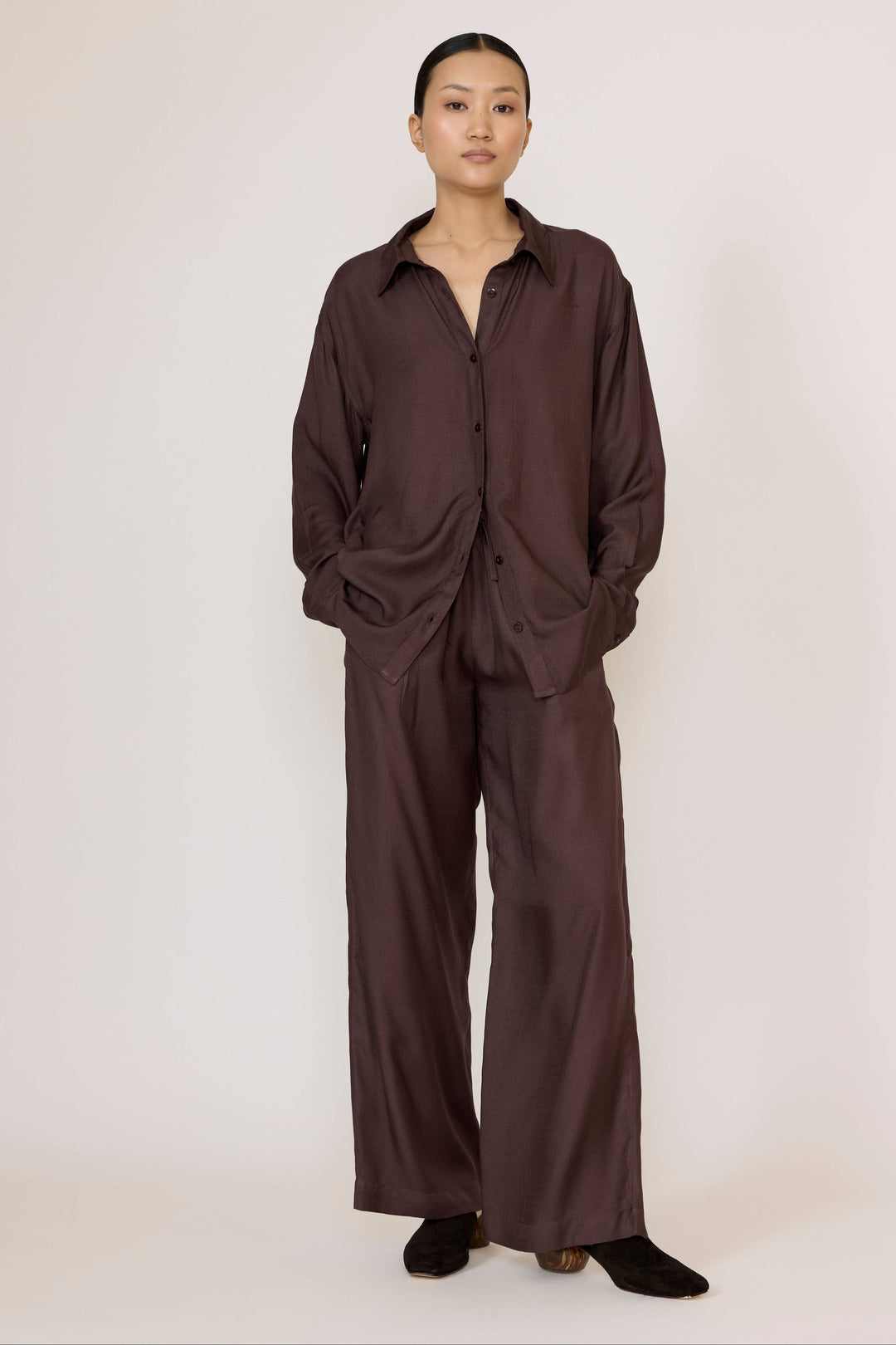 Woman wearing a Laude Everything Shirt in Chocolate Brown with brown pants with a neutral background
