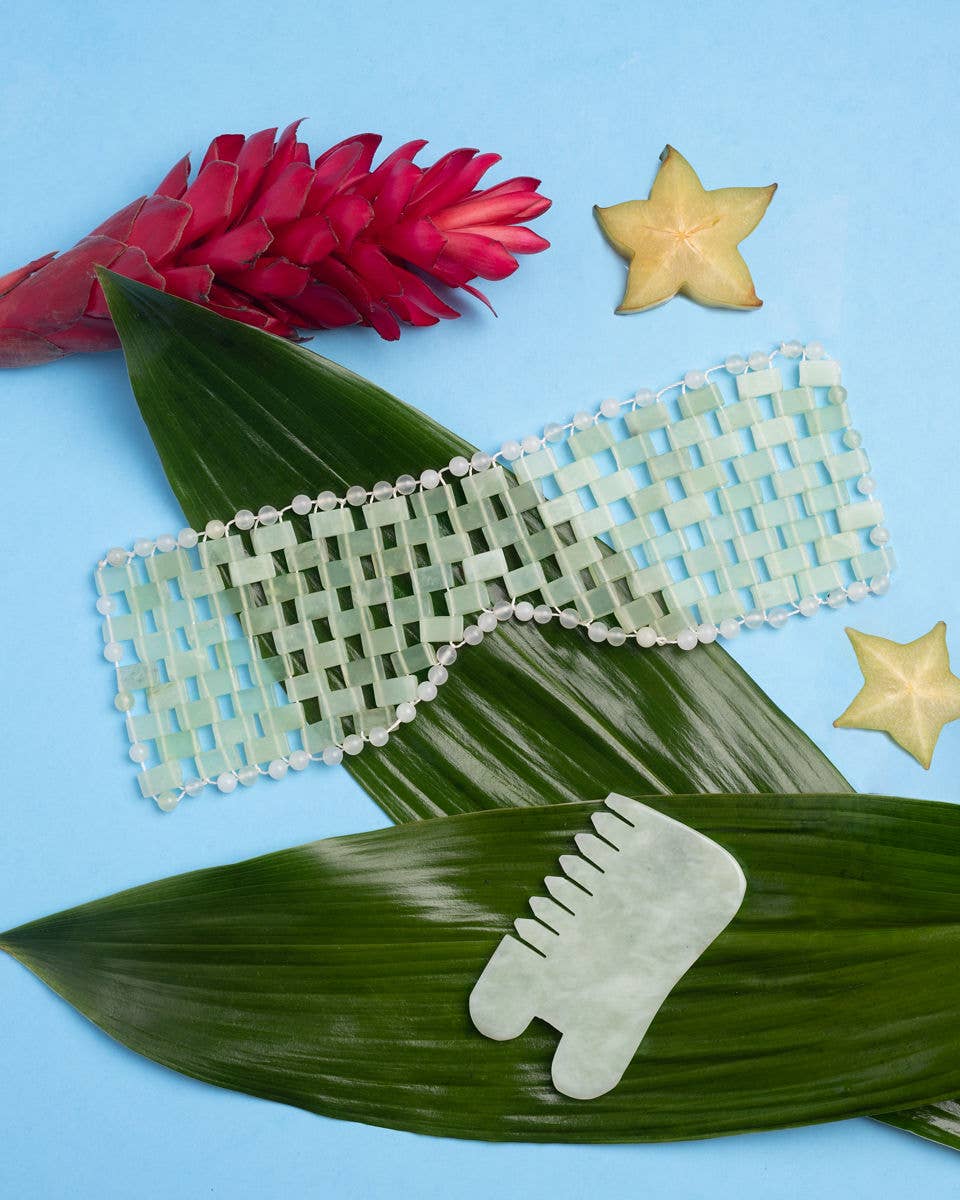  green jade eye mask and gua sha on a blue background with tropical leaves and flowers.