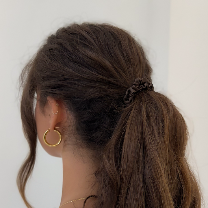 Person with a high ponytail tied back with a brown silk scrunchie wearing gold hoop earrings against a plain background