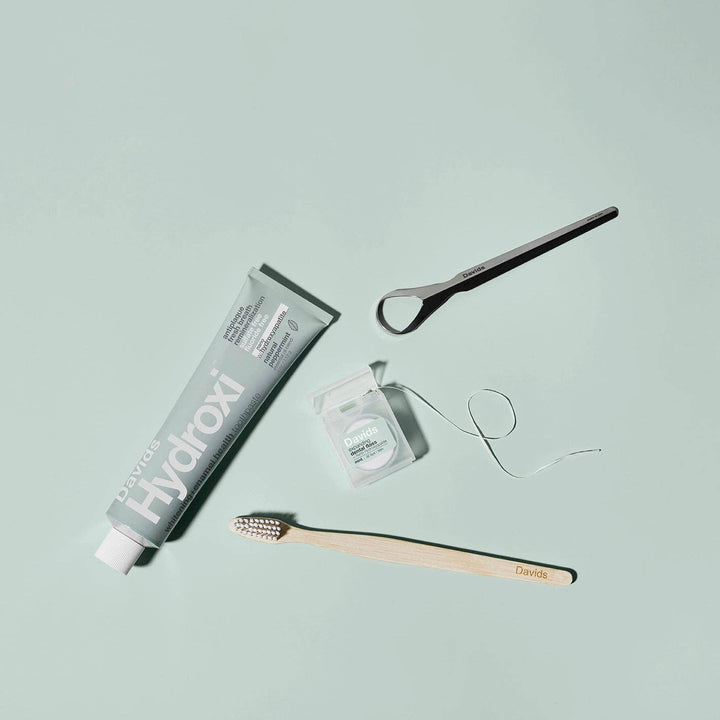 Hydroxi toothpaste tube, wooden toothbrush, dental floss, and small container on a light blue background