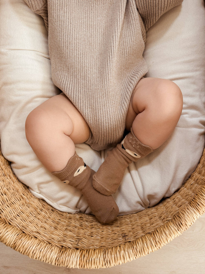 Baby in a beige outfit and brown deer socks lying on a white cushion inside a wicker basket.