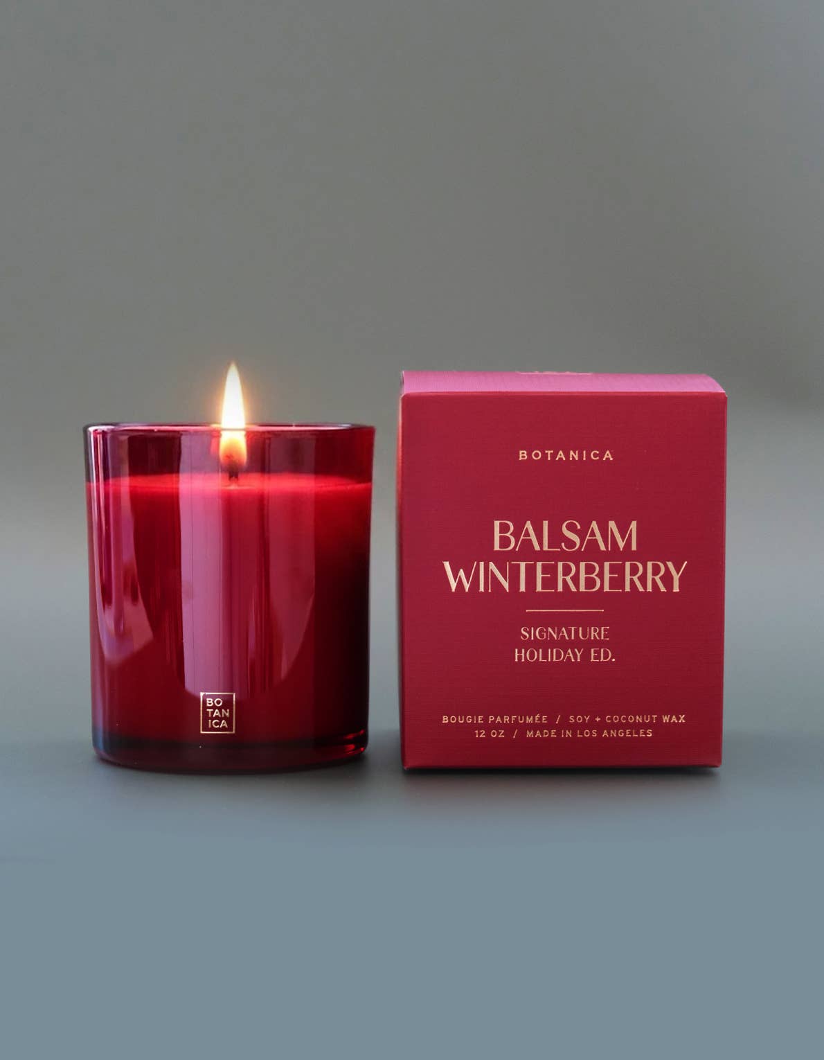 A lit red glass candle jar stands next to its matching red packaging box on a smooth gray background. The box is labeled Botanica Balsam Winterberry.