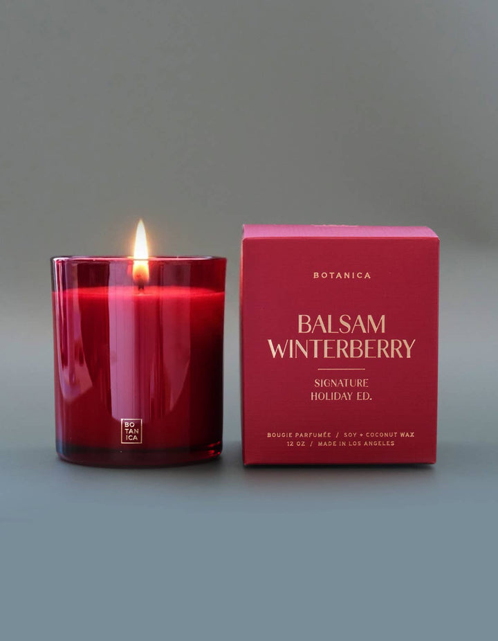A lit red glass candle jar stands next to its matching red packaging box on a smooth gray background. The box is labeled Botanica Balsam Winterberry.
