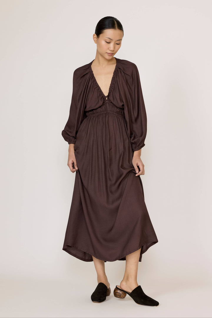 A woman modeling a dark brown midi dress with a deep V-neckline, gathered waist, and long balloon sleeves, paired with black mule shoes against a plain light background.