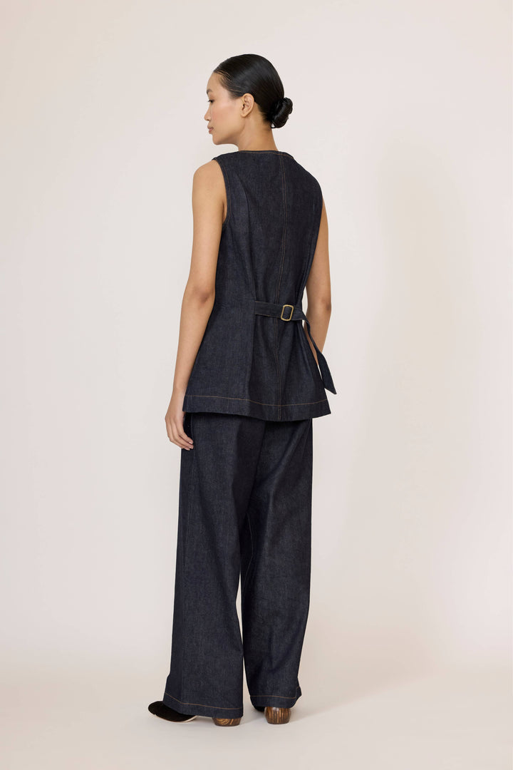 Woman wearing a Laude Longline Vest in Dark Indigo with brown pants over a plain background