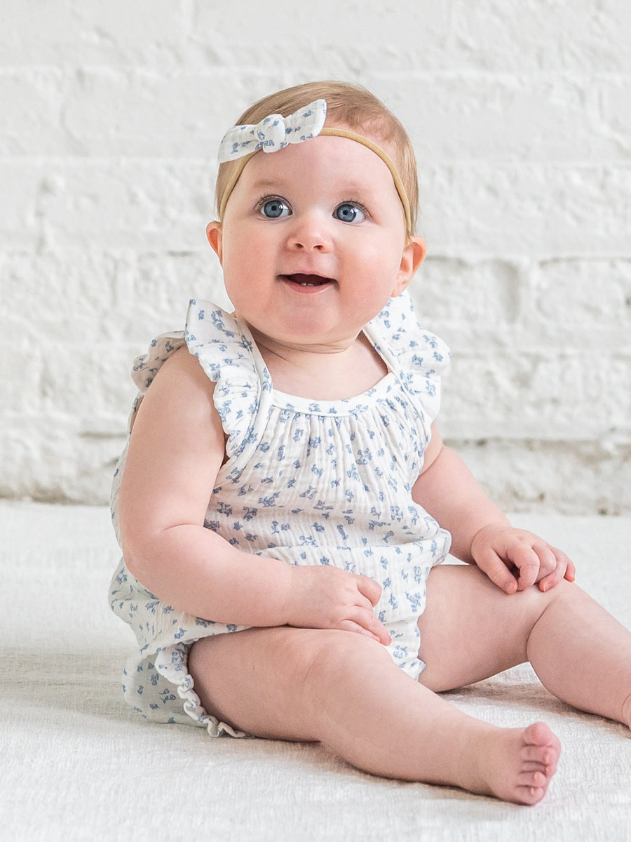 Baby wearing a floral romper and headband sitting on a white surface.