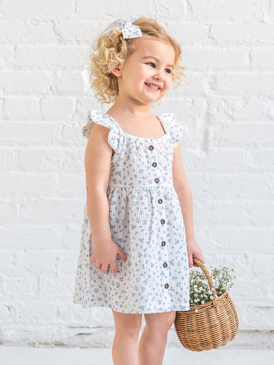 Young girl in a Colored Organics Maisy Ruffle dress holding a basket against a white brick wall