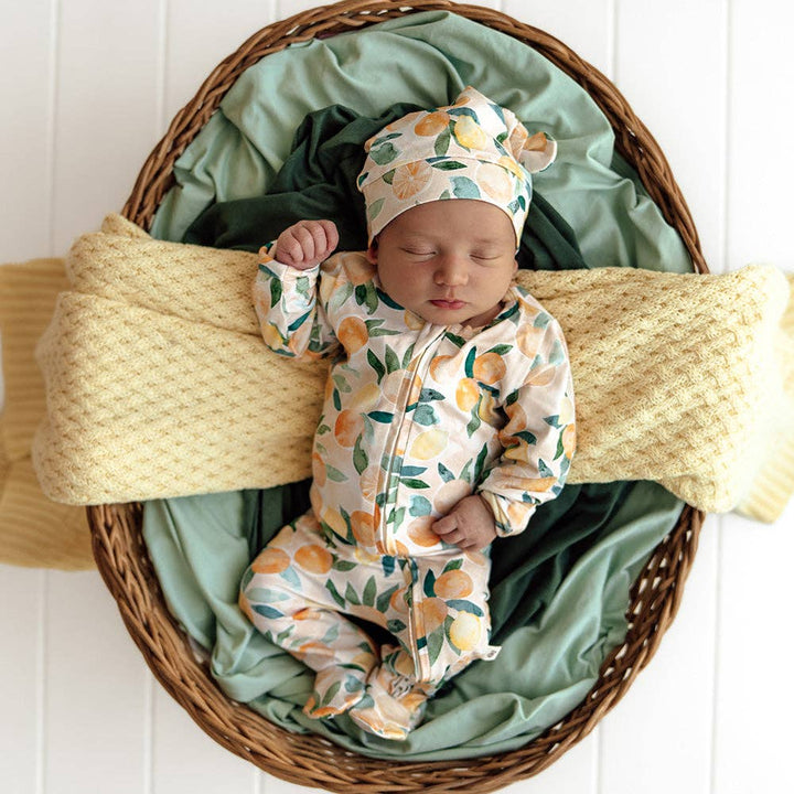 Newborn baby in a Sunny Citrus Organic Snuggle Sleepsuit Zip Footie and hat lying in a woven basket with green and yellow blankets.