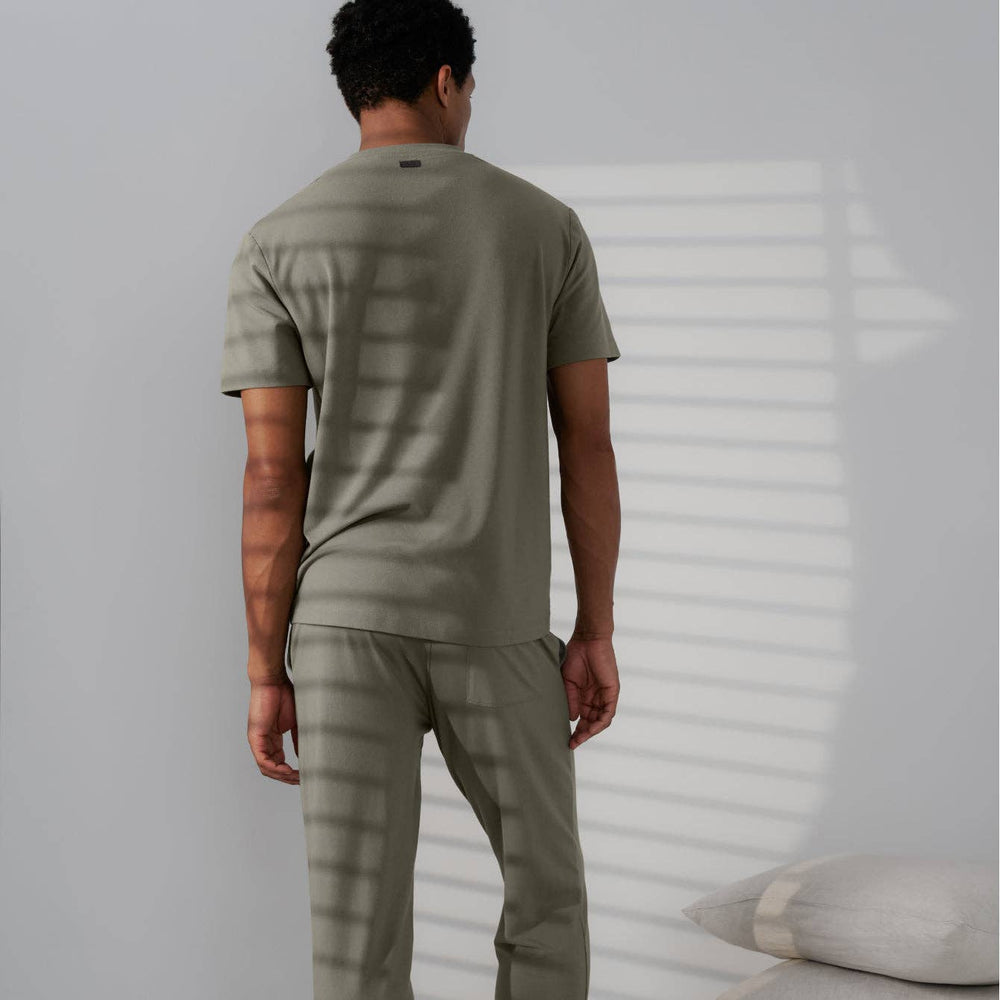 Man wearing a green henley shirt and pants standing against a white wall with a pillow on the floor.