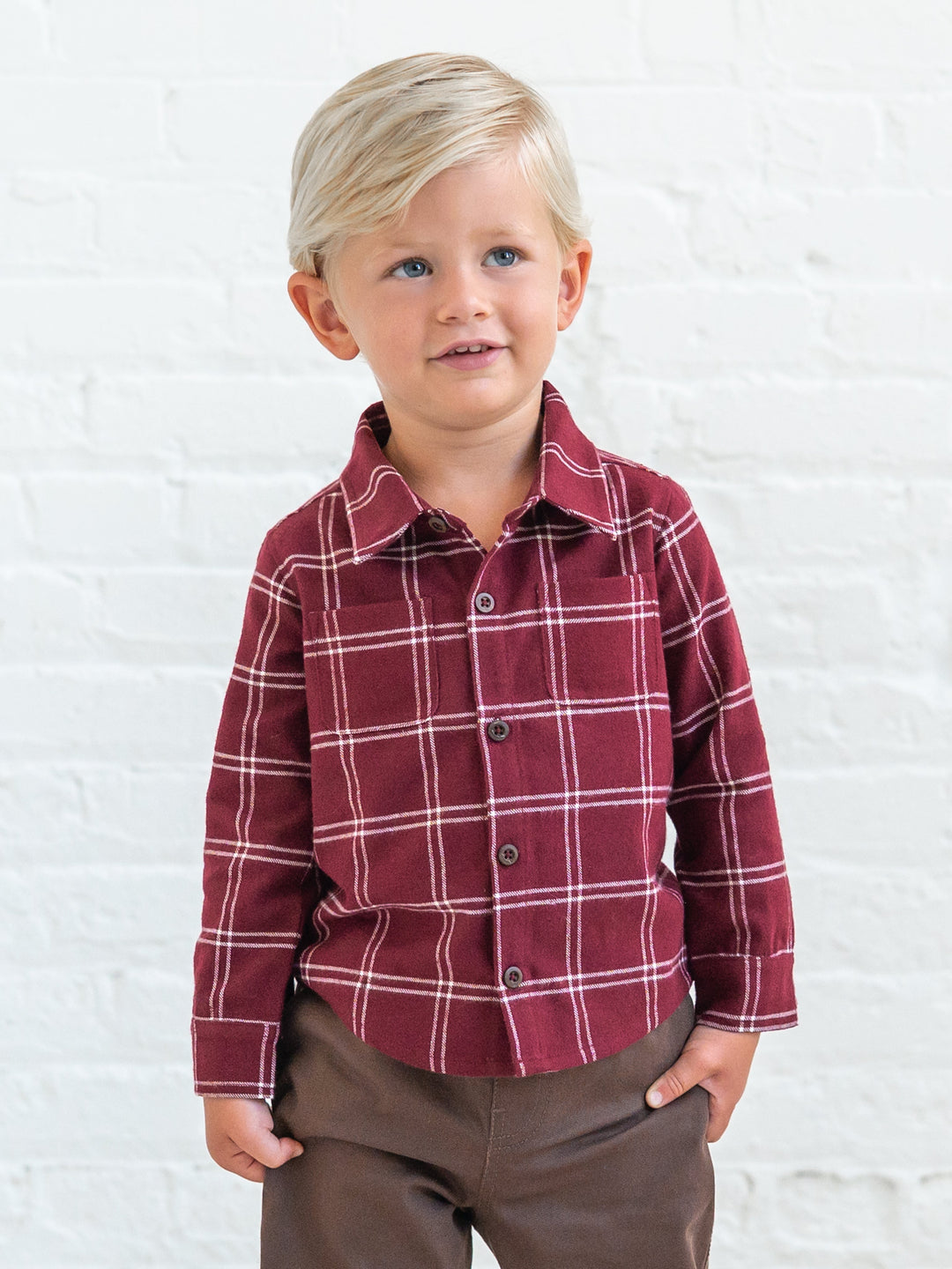 A young blonde boy wearing a Hugo Flannel Long Sleeve Collared Button Down Shirt with brown pants, standing against a white brick wall.