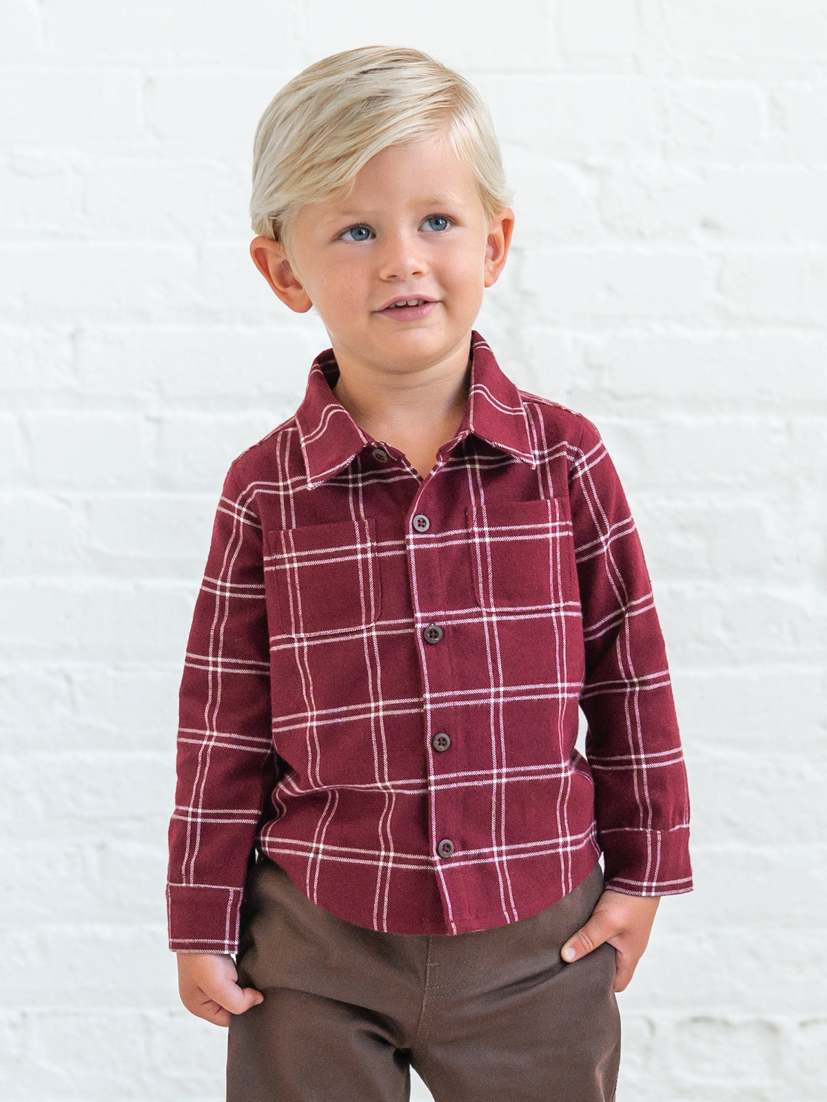 A young blonde boy wearing a Hugo Flannel Long Sleeve Collared Button Down Shirt with brown pants, standing against a white brick wall.