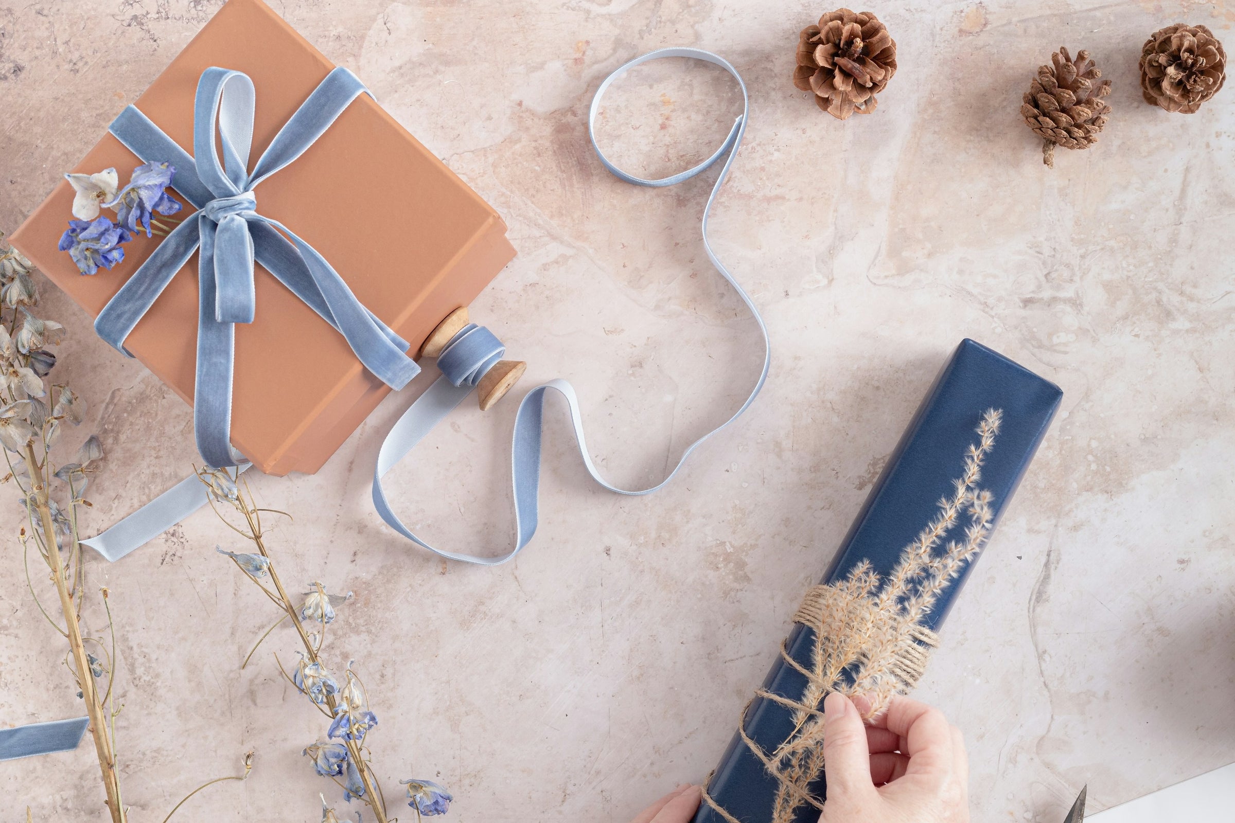 A flatlay image of holiday natural holiday wrapping with blue velvet ribbon, kraft paper boxes, and pine cones