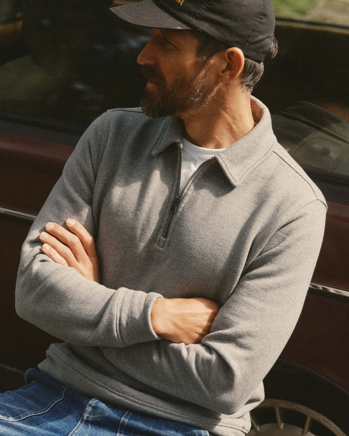 A man wearing a gray quarter-zip long-sleeve pullover sweater, blue jeans, and a black baseball cap, sitting with his arms crossed in front of a dark car.