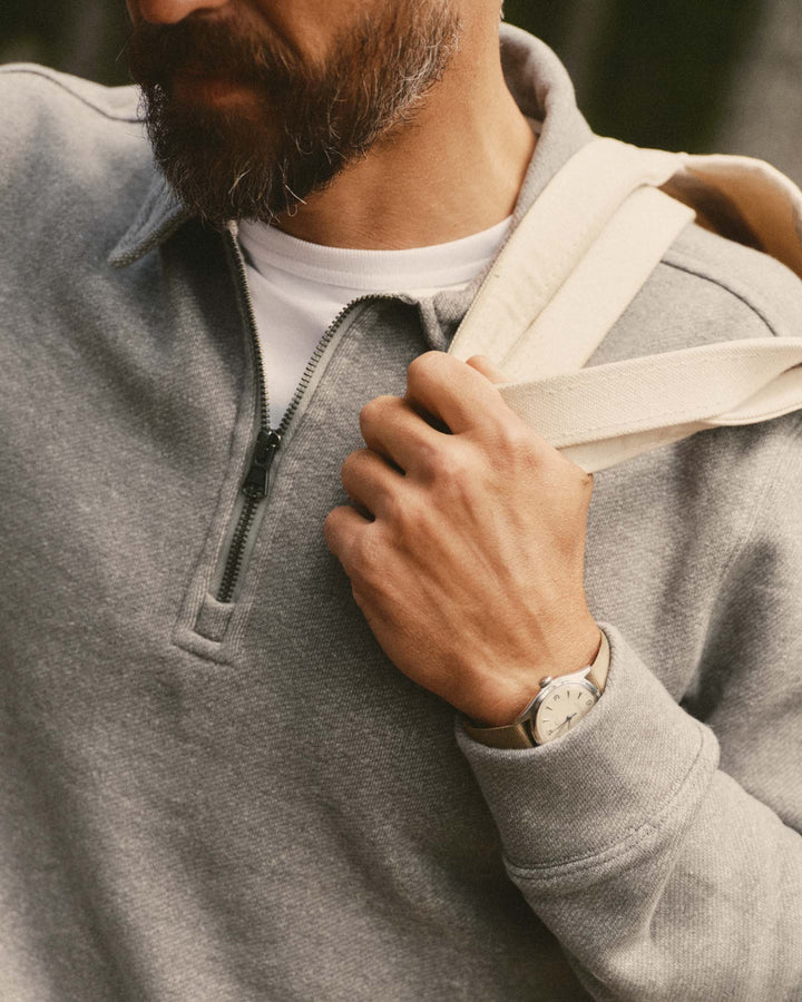 A close-up lifestyle shot of a man wearing a heather gray quarter-zip pullover over a white t-shirt, holding a cream canvas shoulder bag strap over his shoulder.