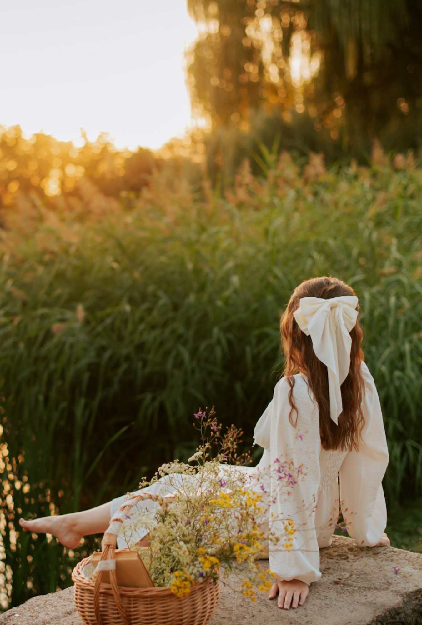 Woman in a white dress sitting on a dock with long grass and wildflowers in the background.