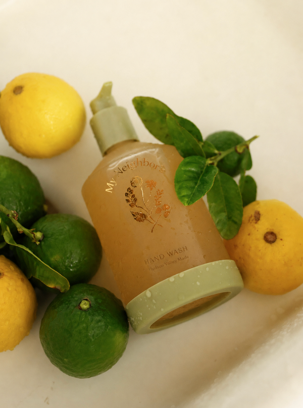 A bottle of My Neighbor's Hand Wash with a light green pump and cap, surrounded by fresh lemons and limes on a white surface.